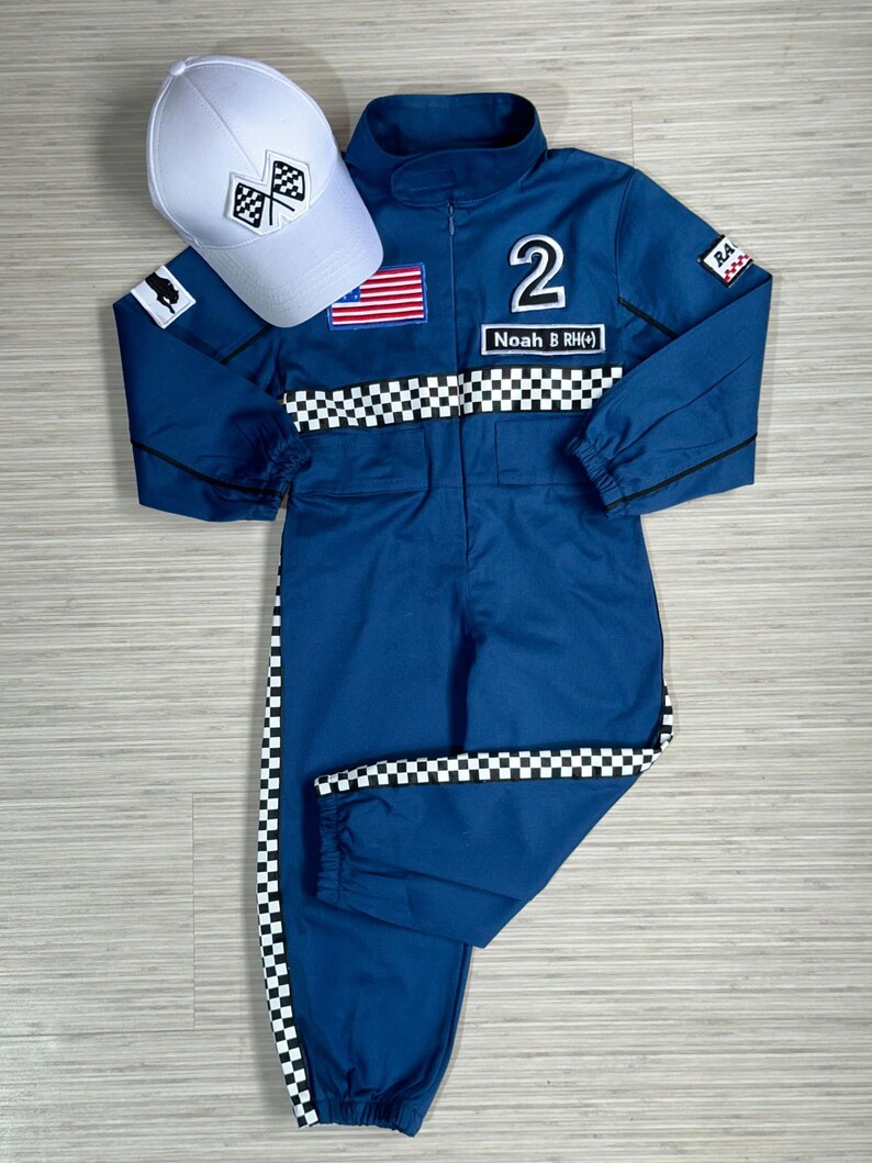 May include: A blue racing jumpsuit with black and white checkered trim. The jumpsuit has a number "2" and the name "Noah B (RH)" on the front. It also has an American flag patch and a black and white checkered flag patch. A white baseball cap with a black and white checkered flag patch is included.