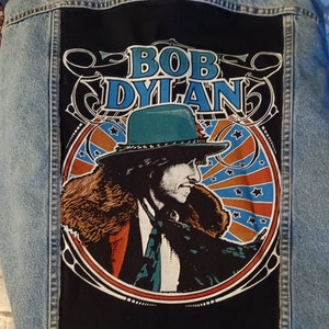 May include: A blue denim jacket with a black patch featuring a vintage illustration of Bob Dylan wearing a hat and fur collar. The patch has the text "BOB DYLAN" in a stylized font.