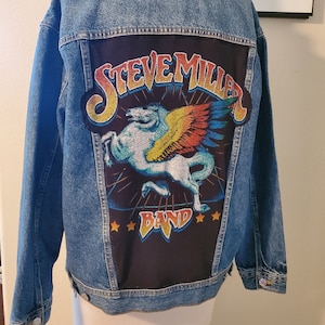 May include: A blue denim jacket with a black patch on the back. The patch features a winged horse with the text "Steve Miller Band" in colorful letters.