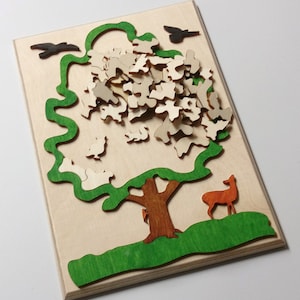 Könnte beinhalten: Ein Holzpuzzle mit einem Baum mit grünem Blätterdach und braunem Stamm. Der Baum ist von einer grünen Grasfläche umgeben. Es gibt mehrere kleine Holzstücke, die in den Baum passen, und ein Reh steht im Gras.