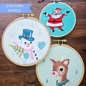 May include: Three Christmas embroidery hoops. One shows Santa Claus in a red suit, another a snowman with a blue top hat, and the third a reindeer with a blue collar. The text "3 PATTERN BUNDLE" is visible.