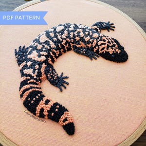 Puede incluir: Diseño bordado de monstruo de Gila sobre tela color melocotón, en un bastidor de bordado de madera. El lagarto presenta patrones negros y color melocotón. El texto en la imagen dice "PDF PATTERN" y "LauraMbrodery".
