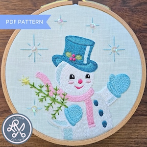 May include: Embroidered snowman design in a wooden hoop. The snowman wears a blue top hat, mittens, and a pink scarf, holding a sprig of greenery. The text "PDF PATTERN" is in the upper left corner. The design is surrounded by embroidered stars.