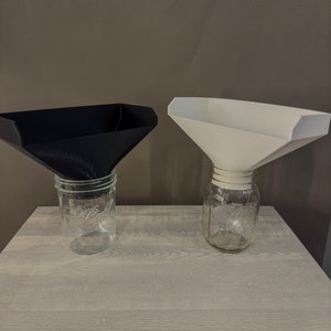 May include: Two plastic funnels, one black and one white, are placed on top of glass jars with lids. The jars are on a light gray wooden surface.
