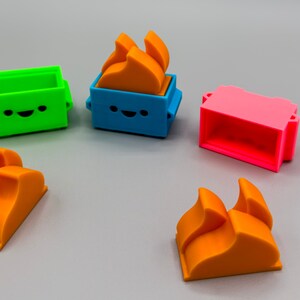 May include: Five colorful 3D printed toy blocks. Two orange blocks are shaped like flames. One green block has a happy face. One blue block has a happy face and an orange flame on top. One pink block has a rectangular opening.