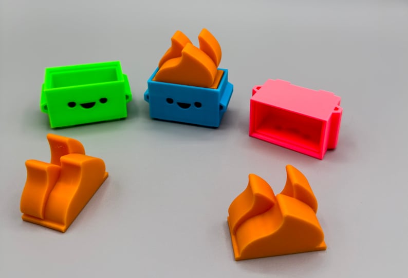 May include: Five colorful 3D printed toy blocks. Two orange blocks are shaped like flames. One green block has a happy face. One blue block has a happy face and an orange flame on top. One pink block has a rectangular opening.