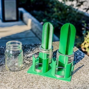 May include: A clear glass mason jar next to a green plastic organizer holding two green cactus-shaped objects. The organizer also holds several silver jar lids. The objects are on a concrete surface.