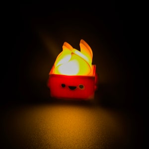 May include: A small, red, box-shaped toy with a smiling face and a yellow flame on top. The toy is lit up from within, casting a warm glow.