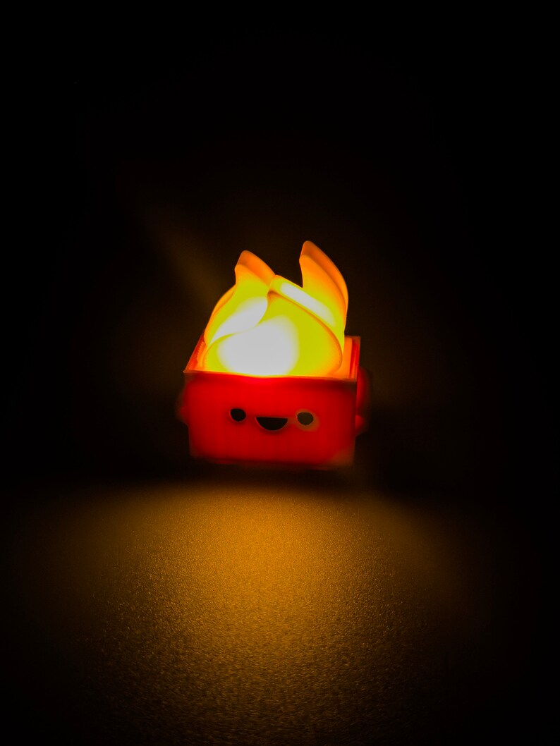 May include: A small, red, box-shaped toy with a smiling face and a yellow flame on top. The toy is lit up from within, casting a warm glow.