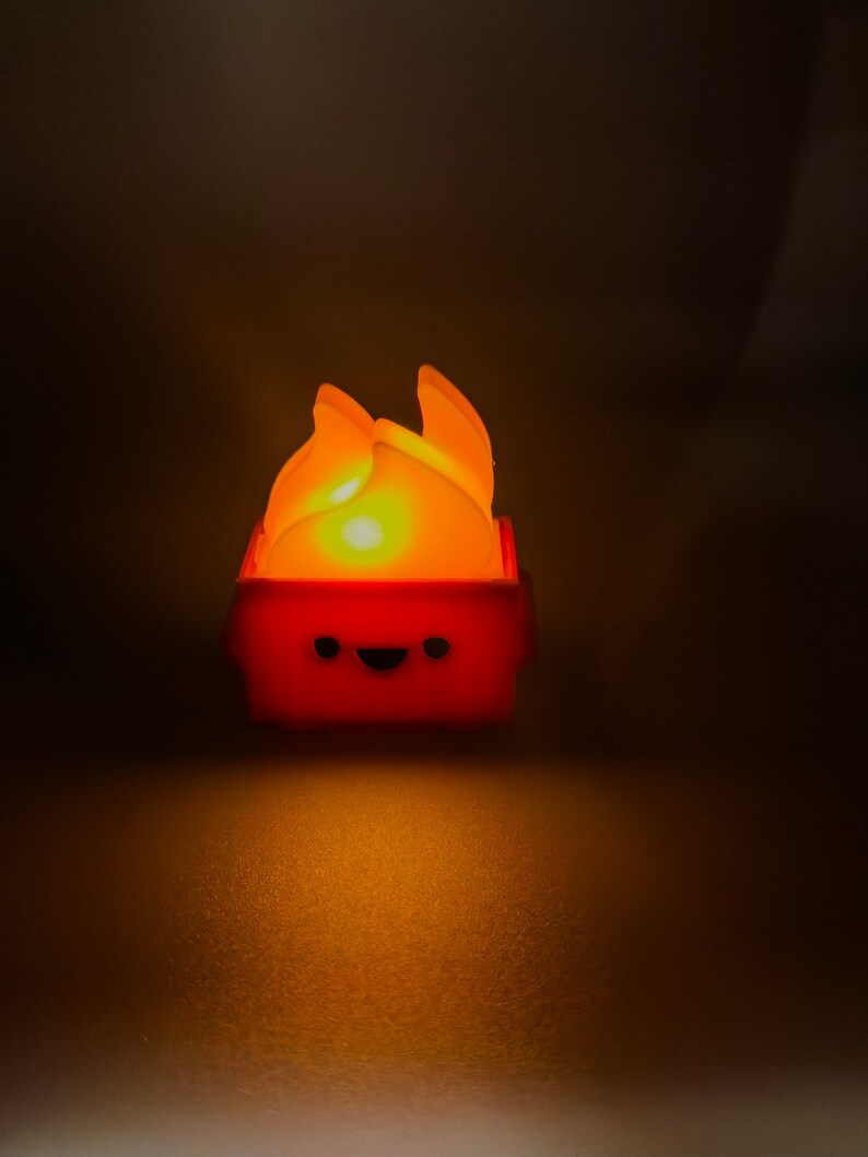 May include: A small, red, fire-shaped nightlight with a happy face. The light is on and glowing orange.