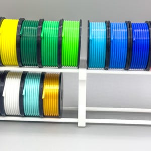 May include: A white plastic spool holder with 14 spools of colorful filament. The filament is arranged in two rows, with seven spools in each row. The colors of the filament are red, orange, yellow, green, lime green, blue, light blue, dark blue, pink, purple, black, white, turquoise, and gold.