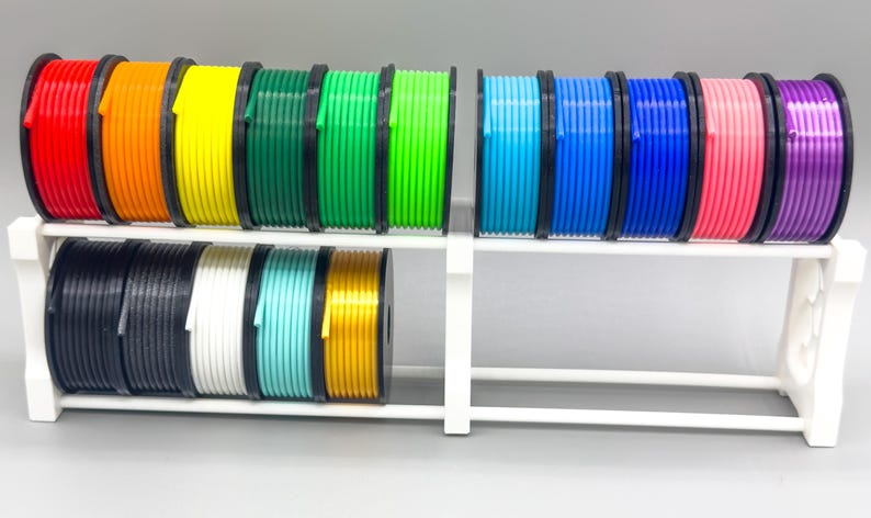 May include: A white plastic spool holder with 14 spools of colorful filament. The filament is arranged in two rows, with seven spools in each row. The colors of the filament are red, orange, yellow, green, lime green, blue, light blue, dark blue, pink, purple, black, white, turquoise, and gold.