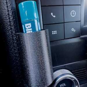 May include: A black plastic car phone holder with a blue and white tube of hand sanitizer inside. The holder is attached to the center console of a car. The console has buttons labeled "MEDIA", "VOL", and icons for phone, clock, and settings.