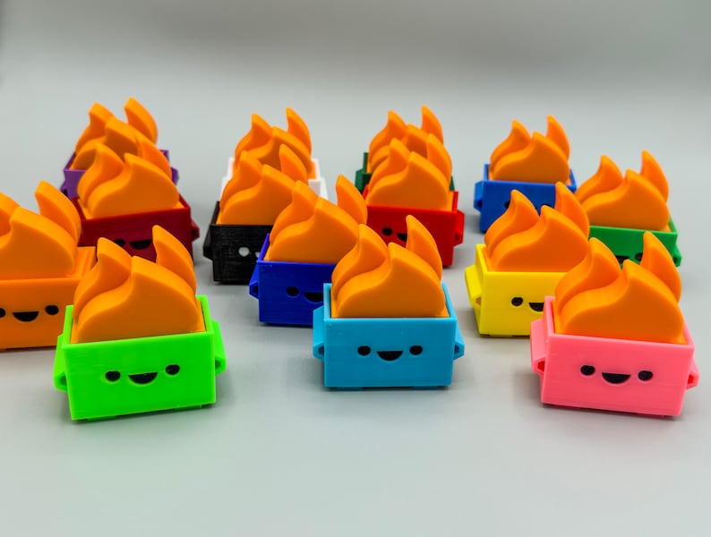 May include: A collection of 12 colorful, small, 3D printed figurines. Each figurine is shaped like a fire with a smiling face and a different colored base. The colors include red, orange, yellow, green, blue, purple, black, and white.