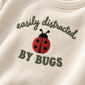 May include: A cream-coloured ribbed bodysuit featuring an embroidered design of a red ladybug with black spots. Above and below the ladybug, the text "easily distracted BY BUGS" is stitched in dark green thread with a textured, raised appearance.