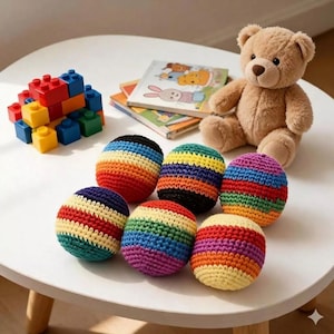 May include: A collection of colorful, crocheted balls with rainbow stripes. The balls are arranged on a white table alongside a teddy bear, building blocks, and children's books. The balls are a mix of red, orange, yellow, green, blue, and purple.