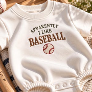 Op de afbeelding: Een crèmekleurige romper met korte mouwen en de geborduurde tekst "APPARENTLY I LIKE BASEBALL" in bruin en groen, met een kleine honkbalafbeelding onder de tekst. Het kledingstuk heeft een geribbelde ronde hals en drukknopen aan de onderkant.