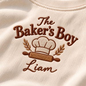 May include: A cream-coloured ribbed sweatshirt featuring brown embroidery that reads The Baker's Boy and Liam. The design includes a central chef hat, a rolling pin, and wheat stalks, all rendered in detailed brown and cream thread stitching.