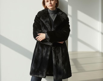 May include: A long, black faux fur coat with a wide collar. The coat is knee-length and has a slightly flared skirt. The model is wearing a gray turtleneck and dark blue jeans. The coat appears to be made of a soft, plush material.