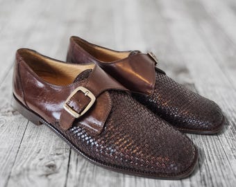 May include: A pair of brown leather dress shoes with woven vamps and buckle fastenings. The shoes have a classic design with a polished finish, suitable for formal occasions. The shoes are displayed on a light grey wooden surface.