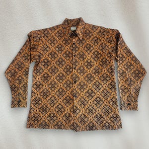 Peut inclure: Chemise à manches longues avec un motif géométrique marron et noir. La chemise a un col et des poignets boutonnés. Le tissu présente un motif répété de diamants et de fleurs. La chemise est boutonnée.