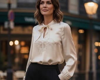 May include: A cream-colored long-sleeve blouse featuring a tie-neck detail and delicate embroidery on the chest, paired with solid black trousers. The blouse has a smooth, satin-like finish with cuffed sleeves and a structured, elegant silhouette.