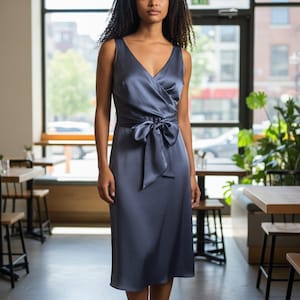 Könnte beinhalten: Ein ärmelloses, knielanges Kleid in einem gedämpften Blau-Grau. Das Kleid hat einen V-Ausschnitt, ein Wickeloberteil und einen passenden Bindegürtel in der Taille. Das Kleid ist aus einem seidigen, fließenden Stoff gefertigt.