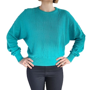 May include: A turquoise blue knitted jumper with a cable knit pattern. The jumper has long sleeves and a round neckline.