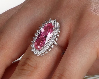 Peut inclure: Une bague en argent ornée d'une grande pierre rose ovale entourée d'un halo de petites pierres claires. La bague est portée sur une main avec des ongles roses, avec des épis de blé en arrière-plan. Le design de la bague est élégant et attrayant.