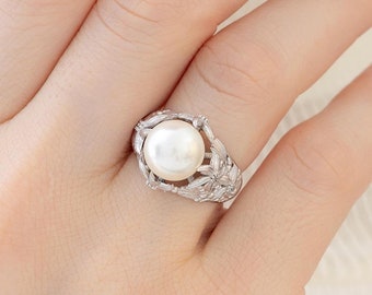 Peut inclure: Une bague argentée ornée d'une perle blanche ronde en son centre. L'anneau en métal présente un motif floral ajouré complexe qui s'étend sur les côtés de la monture, encadrant la perle avec des détails métalliques et de petits accents.