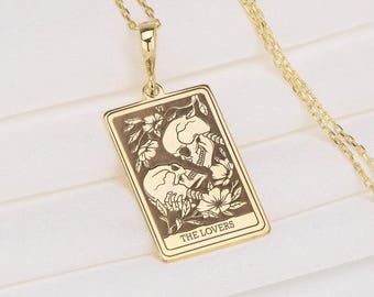 May include: A gold-colored pendant necklace featuring a rectangular pendant with a tarot card design. The card depicts two skulls with floral accents and the text "THE LOVERS". The necklace has a delicate chain.