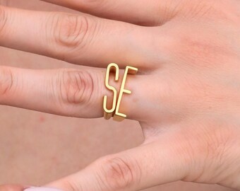 May include: A gold ring displaying the letters "SE" in a connected, minimalist style. The ring is worn on a finger, highlighting its delicate and modern design. The ring is a piece of jewellery.