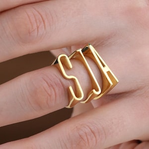 May include: A gold ring displaying the letters "SJA" in a chunky, block-letter design. The ring is worn on a finger, highlighting its personalised style. The ring's metallic lustre and the arrangement of the letters create a contemporary and fashionable accessory.