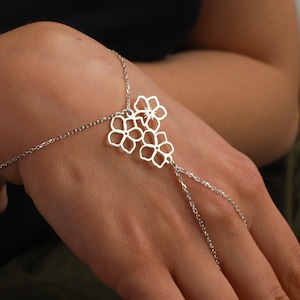 Puede incluir: Un brazalete de cadena de plata con un delicado diseño floral. El brazalete es ajustable y se puede usar en la muñeca o como un anillo de dedo.