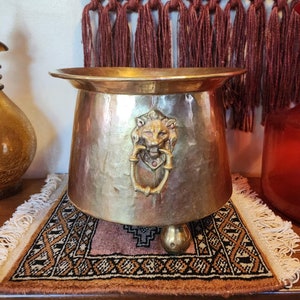 May include: A gold metal planter with a lion head design and three legs. The planter is sitting on a rug with a geometric pattern.