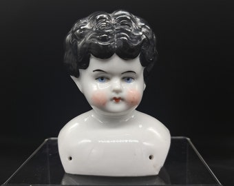 May include: A porcelain doll head with black painted hair, blue eyes, and rosy cheeks. The head is attached to a white porcelain bust.