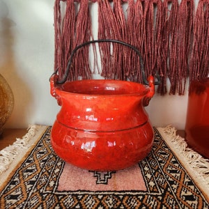 May include: A red ceramic pot with a black metal handle. The pot is sitting on a patterned rug with fringe.