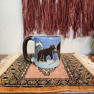 May include: A ceramic mug with a brown handle and a blue body featuring a bear and mountain design. The mug has a light blue interior and sits on a small, patterned rug with a fringed edge. The background includes a red, textured wall hanging.