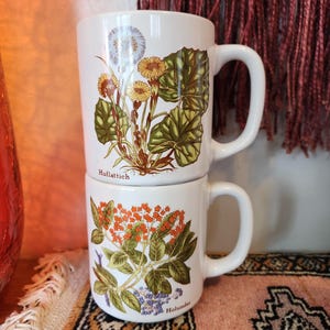 Op de afbeelding: Twee witte keramische mokken met bloemenmotieven. De bovenste mok heeft een witte bloem met groene bladeren en de tekst "Huftlattich". De onderste mok heeft oranje en paarse bloemen met groene bladeren en de tekst "Holunder".