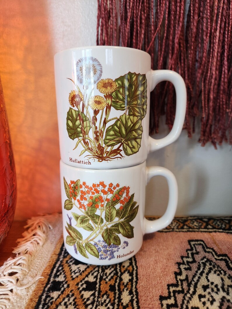 Op de afbeelding: Twee witte keramische mokken met bloemenmotieven. De bovenste mok heeft een witte bloem met groene bladeren en de tekst "Huftlattich". De onderste mok heeft oranje en paarse bloemen met groene bladeren en de tekst "Holunder".