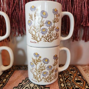 Op de afbeelding: Twee witte keramische mokken met een bloemenpatroon. De mokken hebben een blauw en geel bloemenpatroon met het woord "Kamille" gedrukt op de bodem van elke mok.