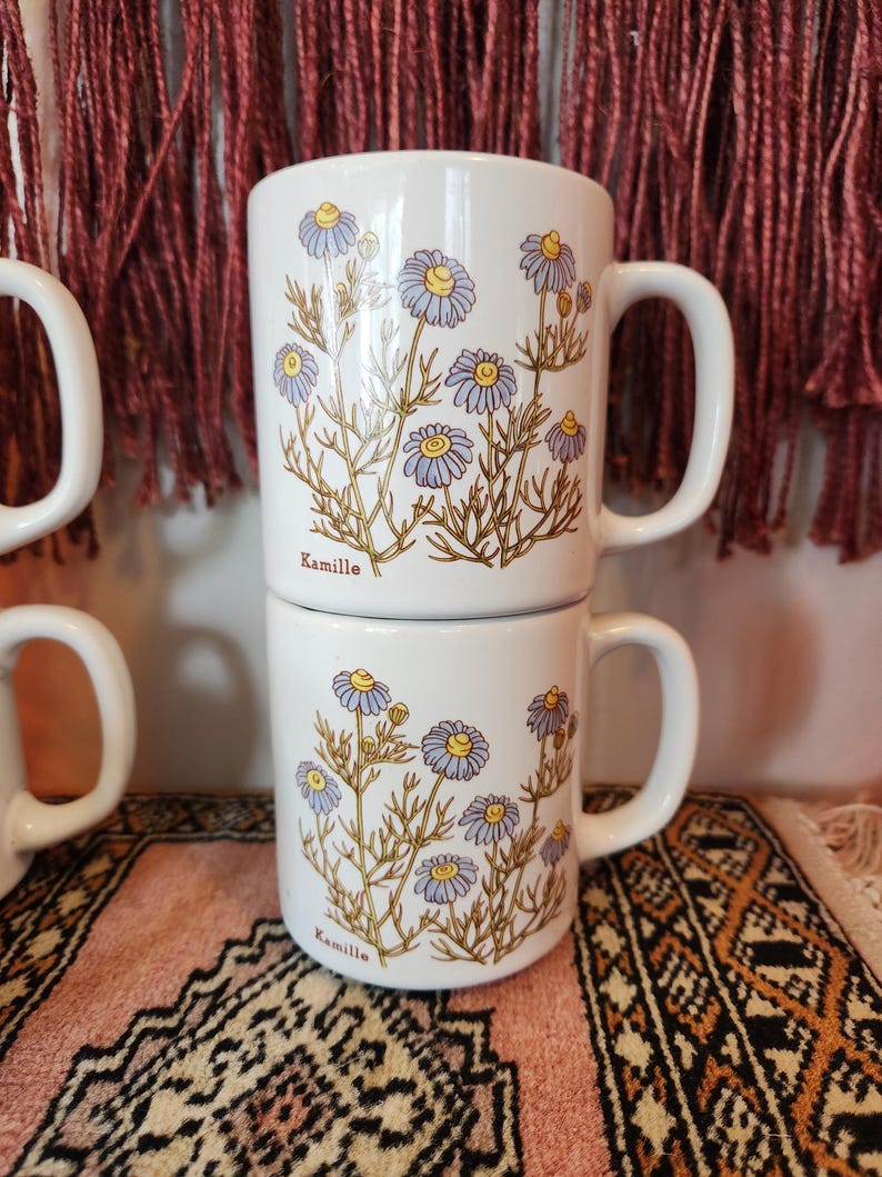 Op de afbeelding: Twee witte keramische mokken met een bloemenpatroon. De mokken hebben een blauw en geel bloemenpatroon met het woord "Kamille" gedrukt op de bodem van elke mok.