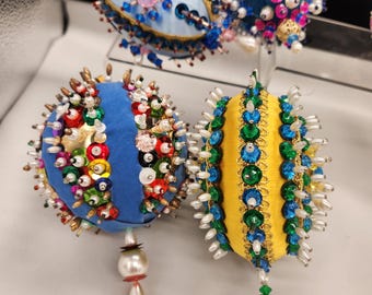 May include: A collection of colorful, handcrafted ornaments. The ornaments feature blue, yellow, and white fabric, adorned with beads, pearls, and decorative accents. Each ornament has a unique shape and design, with dangling pearl-like embellishments.