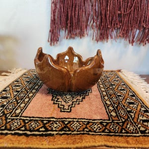 May include: A brown ceramic bowl with a unique petal-like design. The bowl is placed on a small, patterned rug with a pink and orange color scheme. The background features a textured wall and a hanging textile.