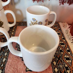 Op de afbeelding: Drie witte keramische mokken met bloemenmotieven. De mokken hebben een eenvoudige, klassieke vorm en zijn perfect voor dagelijks gebruik.