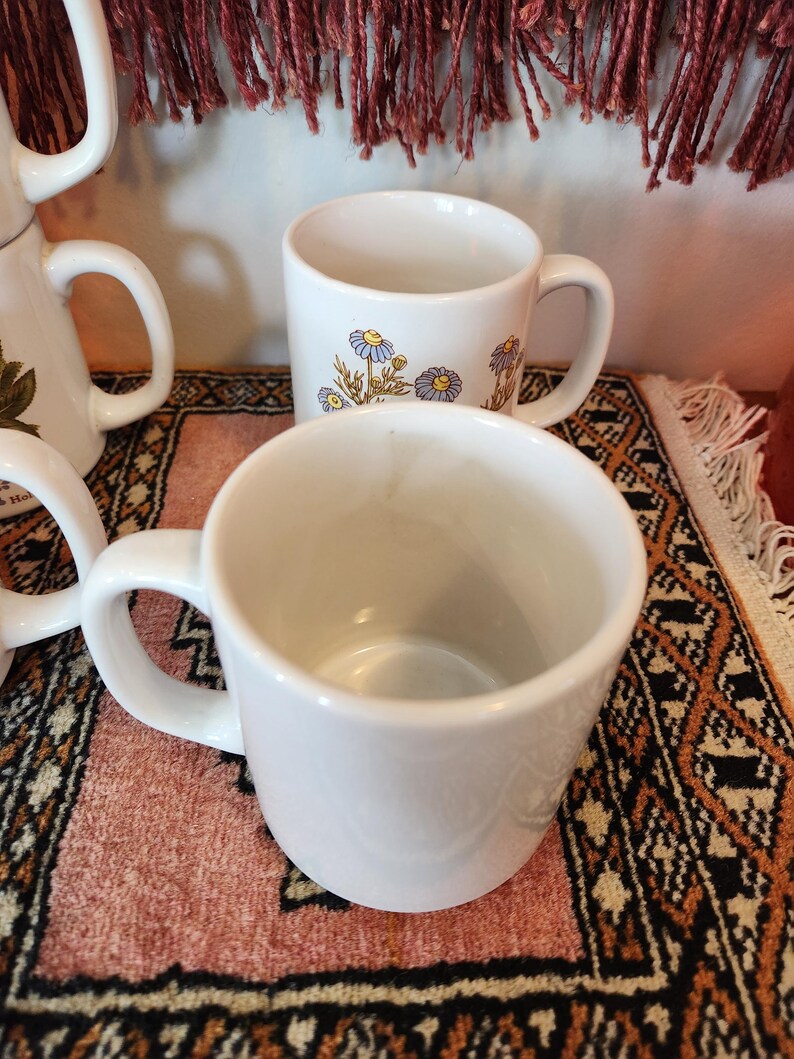Op de afbeelding: Drie witte keramische mokken met bloemenmotieven. De mokken hebben een eenvoudige, klassieke vorm en zijn perfect voor dagelijks gebruik.