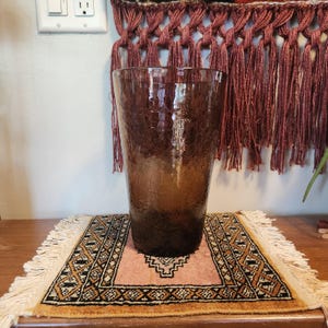May include: A tall, tapered vase made of dark brown glass with a distinct crackle texture throughout its surface. The glass transitions from a lighter amber hue at the top to a deeper, darker brown shade toward the base, featuring a wide opening.