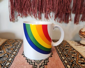 May include: White ceramic mug with a rainbow design in red, orange, yellow, green, and blue. The mug has a curved handle and is set on a pink and black patterned rug. The mug is a standard size.