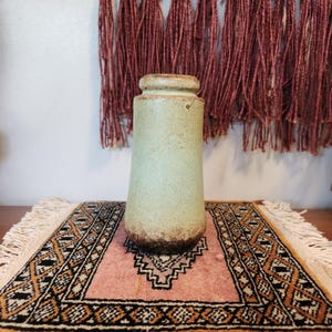 May include: A cylindrical ceramic vase with a tapered neck and a rounded rim. The vase features a speckled light green glaze that transitions into a dark brown, textured finish at the base, showcasing a matte appearance throughout the surface.