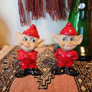 May include: A pair of red and black ceramic elf salt and pepper shakers. The elves have red hats, white faces, and black shoes. They are standing on a patterned rug.